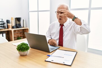 Senior man working at the office using computer laptop peeking in shock covering face and eyes with hand, looking through fingers with embarrassed expression.