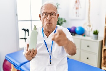 Naklejka na ściany i meble Senior physiotherapy man holding massage body lotion pointing with finger to the camera and to you, confident gesture looking serious