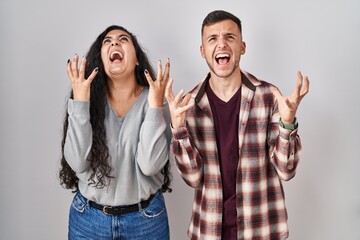 Young hispanic couple standing over white background crazy and mad shouting and yelling with aggressive expression and arms raised. frustration concept.