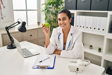 Young hispanic woman wearing dietitian uniform holding apple at clinic