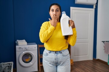 Young hispanic woman holding detergent bottle covering mouth with hand, shocked and afraid for...