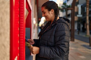 Young hispanic woman holding money of automatic teller bank machine at street