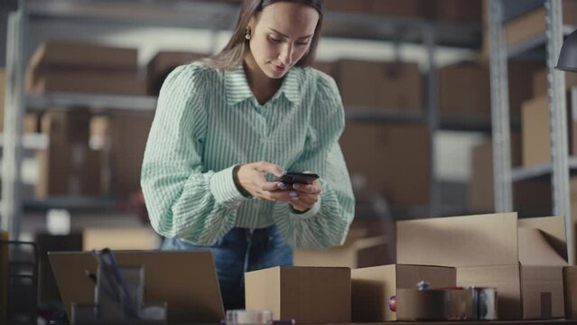 Inventory Manager Using Smartphone to Scan a Barcode on Parcel, Preparing a Small Cardboard Box for Postage. Young Female Small Business Owner Working on Laptop in Warehouse with Colleague.