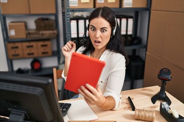 Young hispanic woman working at small business speaking on video call scared and amazed with open mouth for surprise, disbelief face