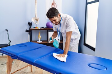 Young hispanic man wearing physiotherapist uniform disinfecting massage table at rehab clinic