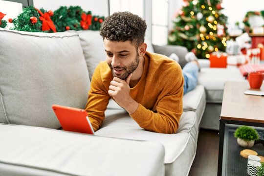 Young arab man using touchpad lying on the sofa by christmas tree at home.