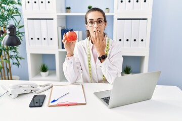 Young hispanic woman working dietitian clinic covering mouth with hand, shocked and afraid for...