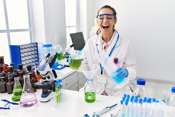Fototapeta premium Young hispanic woman working at scientist laboratory celebrating crazy and amazed for success with open eyes screaming excited.