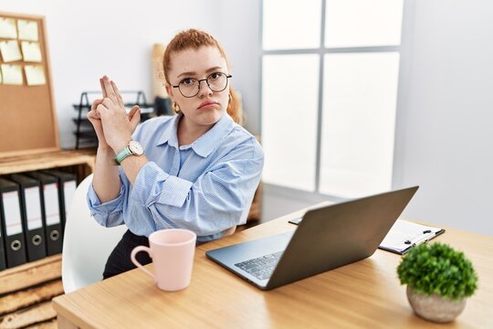 Young redhead woman working at the office using computer laptop holding symbolic gun with hand gesture, playing killing shooting weapons, angry face