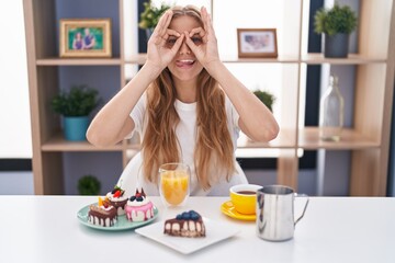 Obraz na płótnie Canvas Young caucasian woman eating pastries t for breakfast doing ok gesture like binoculars sticking tongue out, eyes looking through fingers. crazy expression.