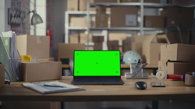 Laptop Computer Standing on a Table with a Green Screen Chromakey Mock Up Display. Small Business Warehouse with Cardboard Boxes in the Background. Work Desk with Parcels. Zoom In Shot.