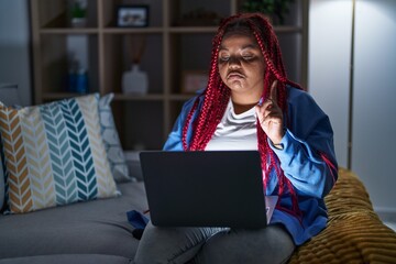 African american woman with braided hair using computer laptop at night pointing up looking sad and...