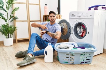 Young hispanic man putting dirty laundry into washing machine in hurry pointing to watch time, impatience, upset and angry for deadline delay