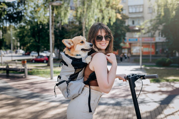 Happy smiling woman traveler is riding her electro scooter in city parkland with dog Welsh Corgi Pembroke in a special backpack