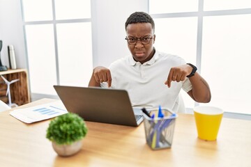 Young african man working at the office using computer laptop pointing down looking sad and upset,...