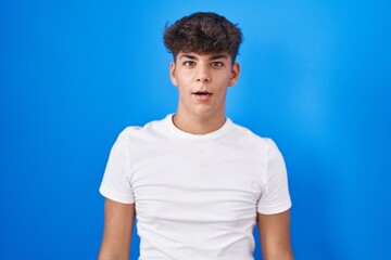 Hispanic teenager standing over blue background afraid and shocked with surprise and amazed expression, fear and excited face.