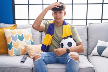 Hispanic teenager sitting on the sofa watching football match stressed and frustrated with hand on...