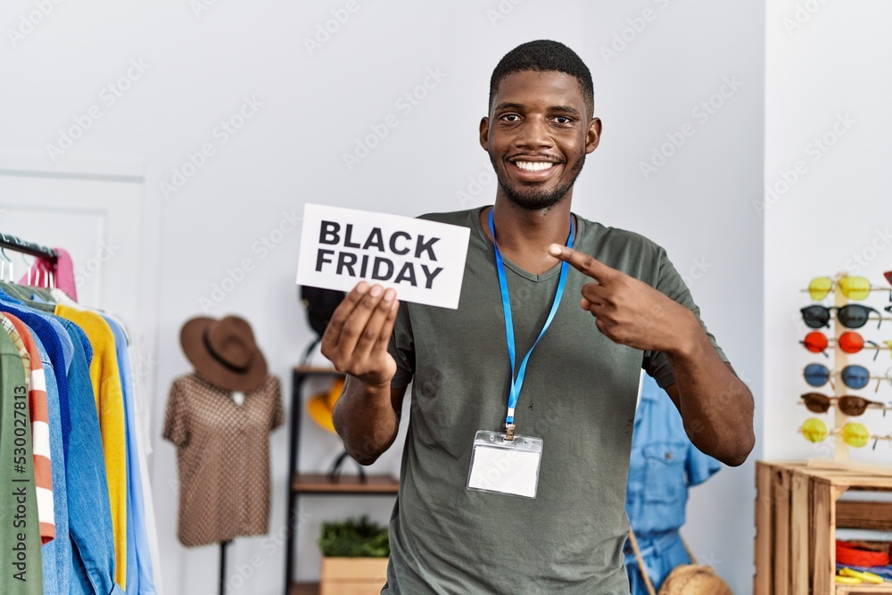 Sticker Young african american man holding black friday banner at retail shop smiling happy pointing with hand and finger