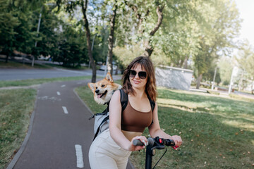 Fototapeta na wymiar Happy smiling woman traveler is riding her electro scooter in city parkland with dog Welsh Corgi Pembroke in a special backpack