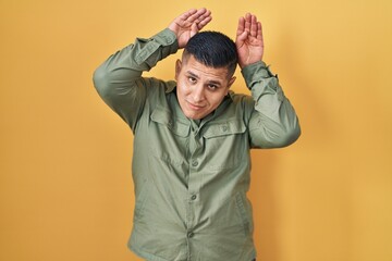 Hispanic young man standing over yellow background doing bunny ears gesture with hands palms...