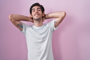 Young hispanic man standing over pink background relaxing and stretching, arms and hands behind...