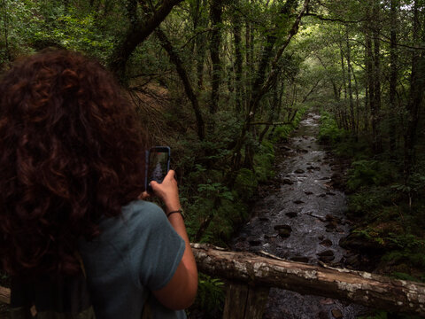 Brunette woman taking a photo on a wood bridge with river flowing through an oak and chestnut trees in the background.