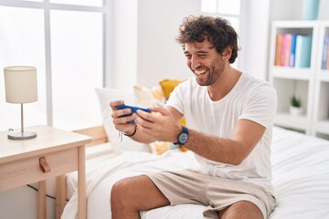 Young hispanic man watching video on smartphone sitting on bed at bedroom