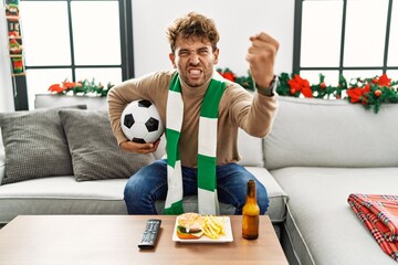 Young hispanic man football hooligan holding ball at home annoyed and frustrated shouting with...