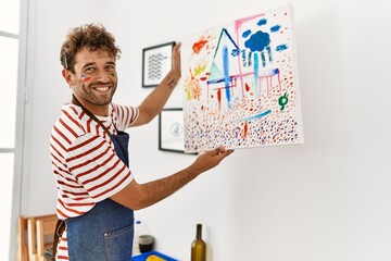 Young hispanic man smiling confident hanging canvas on wall at art studio