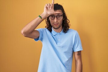 Young hispanic man standing over yellow background making fun of people with fingers on forehead...