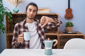 Young hispanic man drinking coffee from french coffee maker with angry face, negative sign showing...