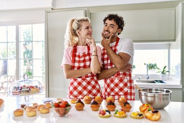 Couple of wife and husband cooking pastries at the kitchen looking confident at the camera smiling with crossed arms and hand raised on chin. thinking positive.