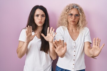 Mother and daughter standing together over pink background moving away hands palms showing refusal and denial with afraid and disgusting expression. stop and forbidden.