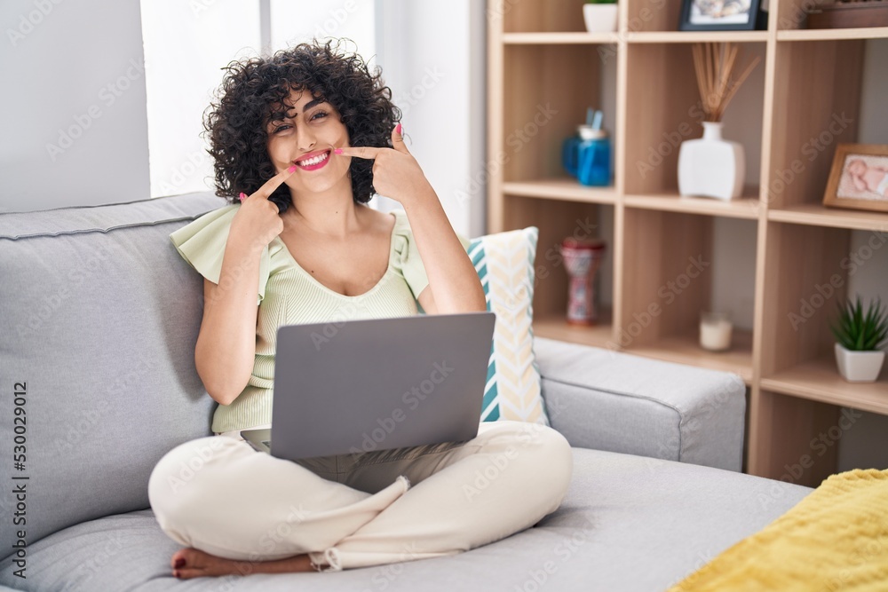 Poster Young brunette woman with curly hair using laptop sitting on the sofa at home smiling cheerful showing and pointing with fingers teeth and mouth. dental health concept.