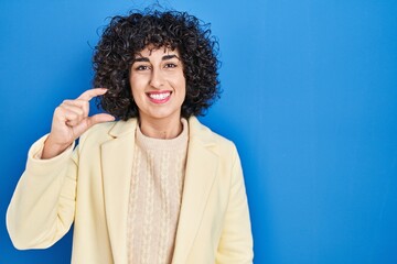 Fototapeta premium Young brunette woman with curly hair standing over blue background smiling and confident gesturing with hand doing small size sign with fingers looking and the camera. measure concept.