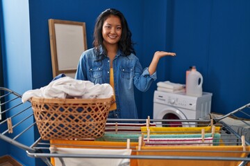 Young asian woman hanging clothes at clothesline smiling cheerful presenting and pointing with palm of hand looking at the camera.