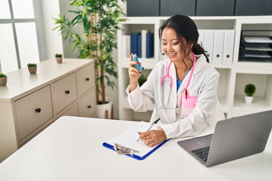 Young chinese woman wearing doctor uniform holding inhaler writing on document at clinic