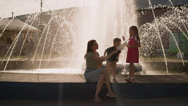 Little boy bites cotton candy while mother holds stick and sister stands nearby on fountain parapet slow motion. Friendly family shares dessert in park
