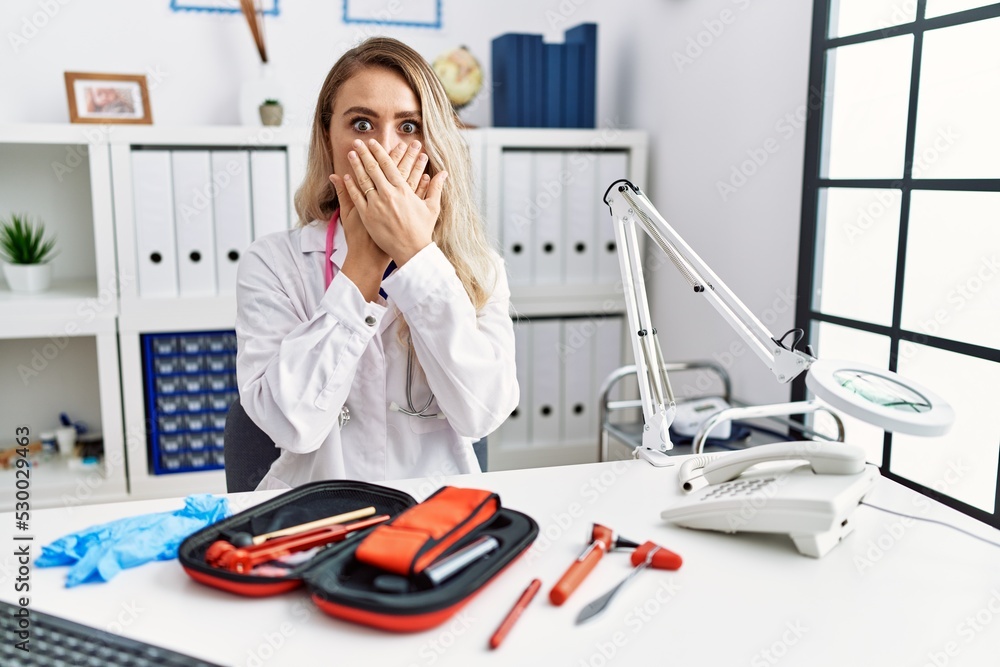 Poster young beautiful doctor woman with reflex hammer and medical instruments shocked covering mouth with 