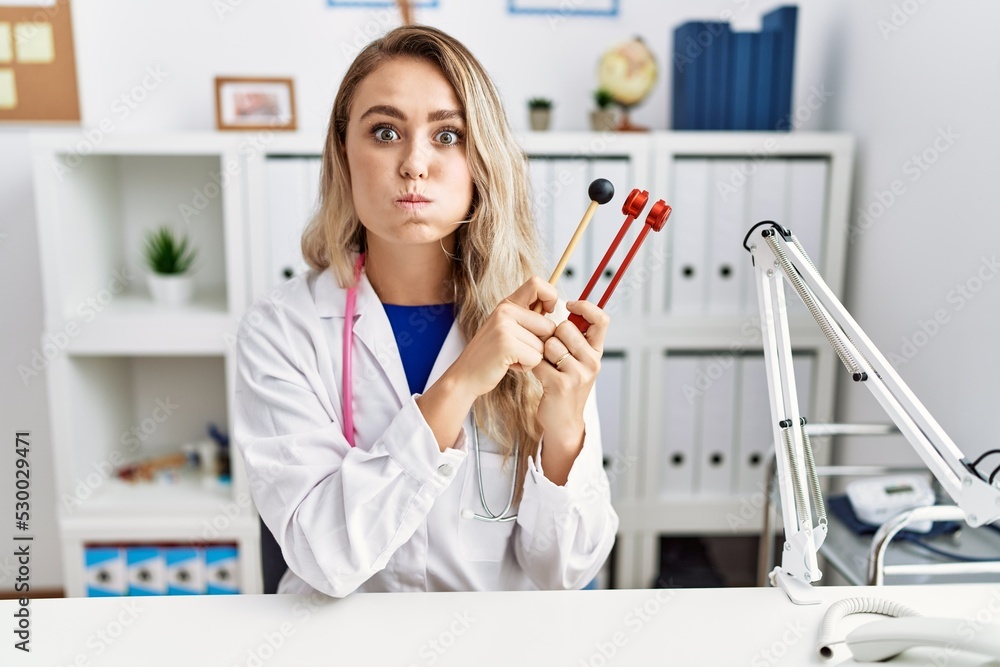 Sticker Young beautiful doctor woman holding diapason instrument puffing cheeks with funny face. mouth inflated with air, catching air.