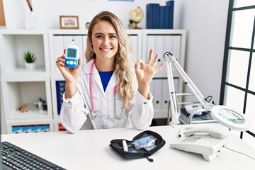 Young beautiful doctor woman holding glucose meter doing ok sign with fingers, smiling friendly gesturing excellent symbol