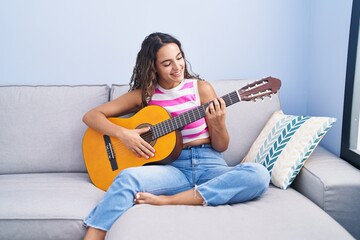 Young beautiful hispanic woman playing classical guitar sitting on sofa at home