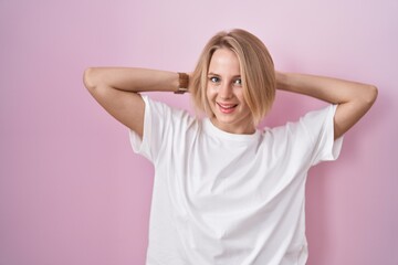 Young caucasian woman standing over pink background relaxing and stretching, arms and hands behind head and neck smiling happy