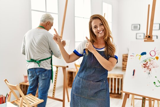 Hispanic woman wearing apron at art studio smiling and looking at the camera pointing with two hands and fingers to the side.