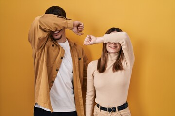 Young hispanic couple standing over yellow background covering eyes with arm smiling cheerful and...