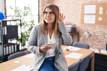 Young hispanic woman working at the office wearing glasses smiling with hand over ear listening an hearing to rumor or gossip. deafness concept.