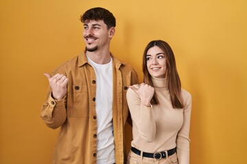Young hispanic couple standing over yellow background smiling with happy face looking and pointing to the side with thumb up.
