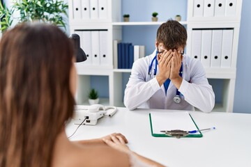 Young doctor with client at medical clinic with sad expression covering face with hands while...