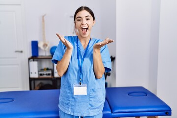 Obraz premium Young hispanic woman wearing physiotherapist uniform standing at clinic celebrating crazy and amazed for success with arms raised and open eyes screaming excited. winner concept
