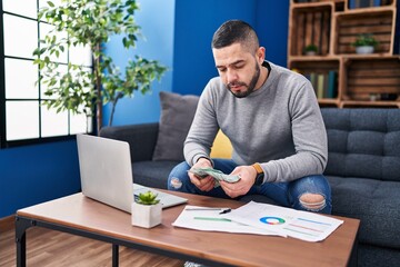 Young latin man using laptop holding dollars sitting on table at home